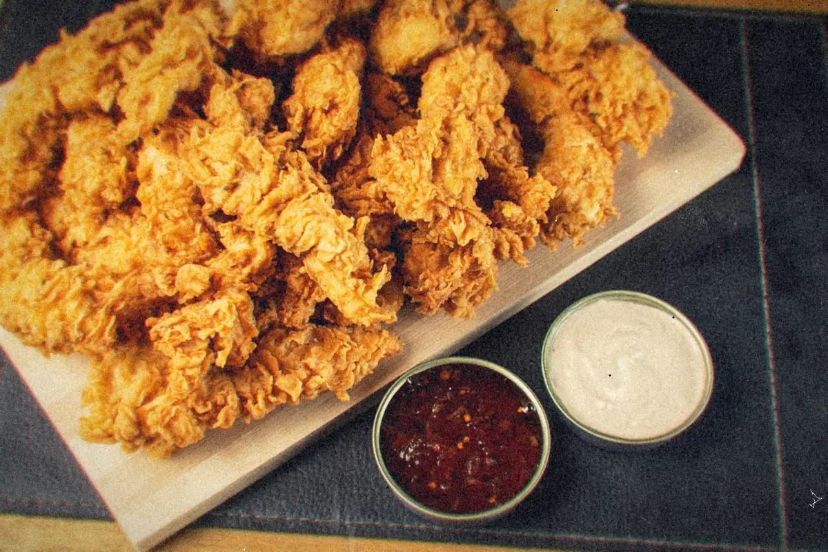 A board piled with crispy golden chicken tenders next to two small dipping sauces.