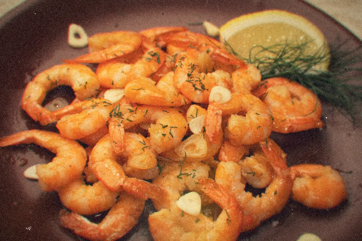 A plate of glossy garlic shrimp with sliced garlic, herbs, a lemon wedge and dill.
