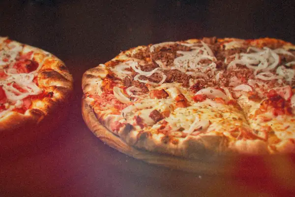 Two blistered-crust pizzas topped with meat, cheese and sliced onions on a wooden table.