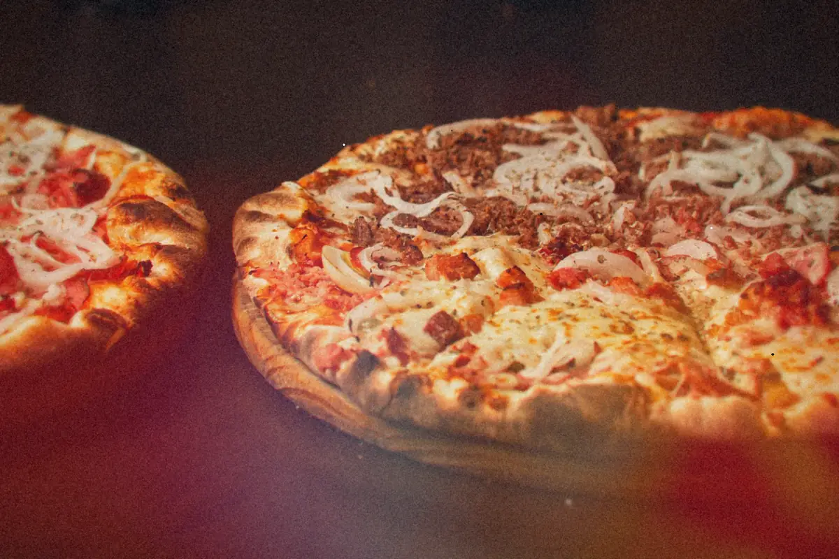 Two blistered-crust pizzas topped with meat, cheese and sliced onions on a wooden table.