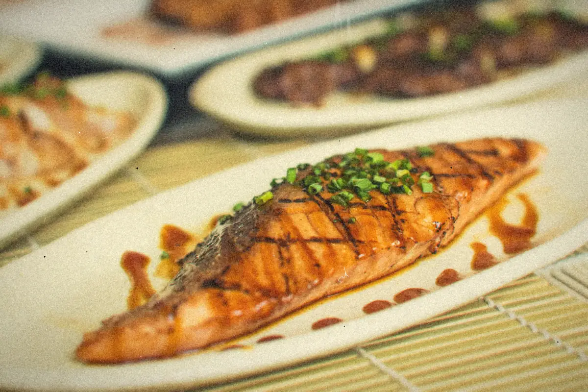 A grilled salmon fillet with a dark soy glaze and sliced green onions on a white plate.