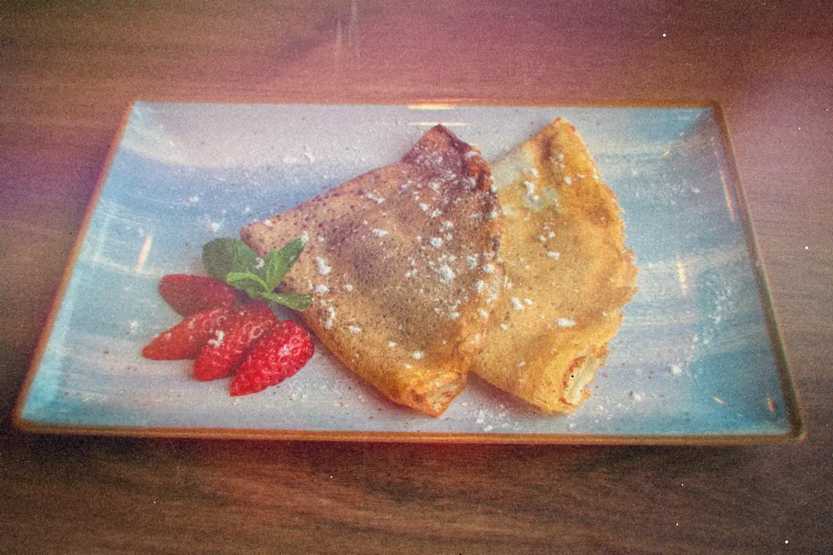 Two folded crepes dusted with powdered sugar on a blue plate with sliced strawberries and mint.