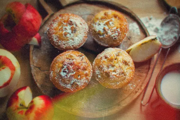 Four sugar-dusted apple muffins on a wooden board with sliced red apples.