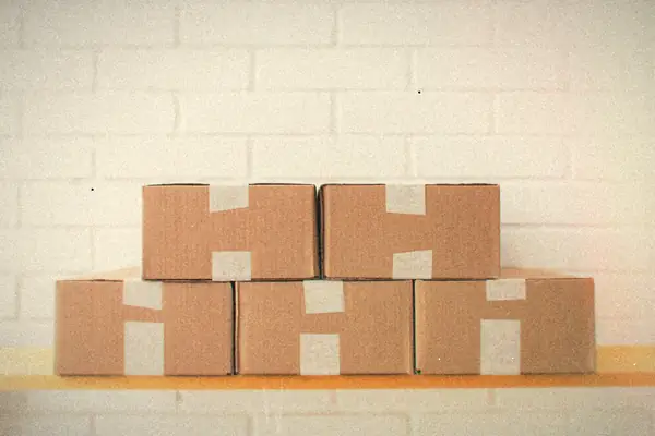 A neat stack of taped cardboard boxes on a wooden shelf against a white brick wall.