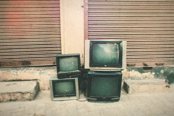 Old CRT televisions and monitors left out on a sidewalk in front of a shuttered storefront.