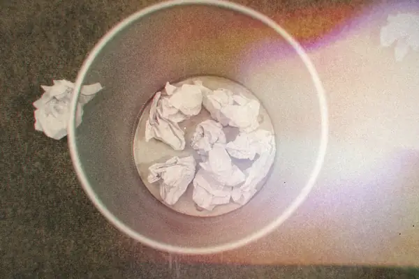 A wire wastebasket full of crumpled paper on a carpeted floor.