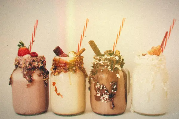 Four loaded milkshakes in mason jars with striped straws, topped with chocolate, cookies, wafers and a strawberry.