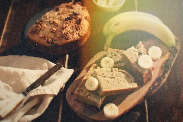 Pieces of banana bread with banana slices on a wooden board, a whole banana and a walnut-topped loaf behind them.