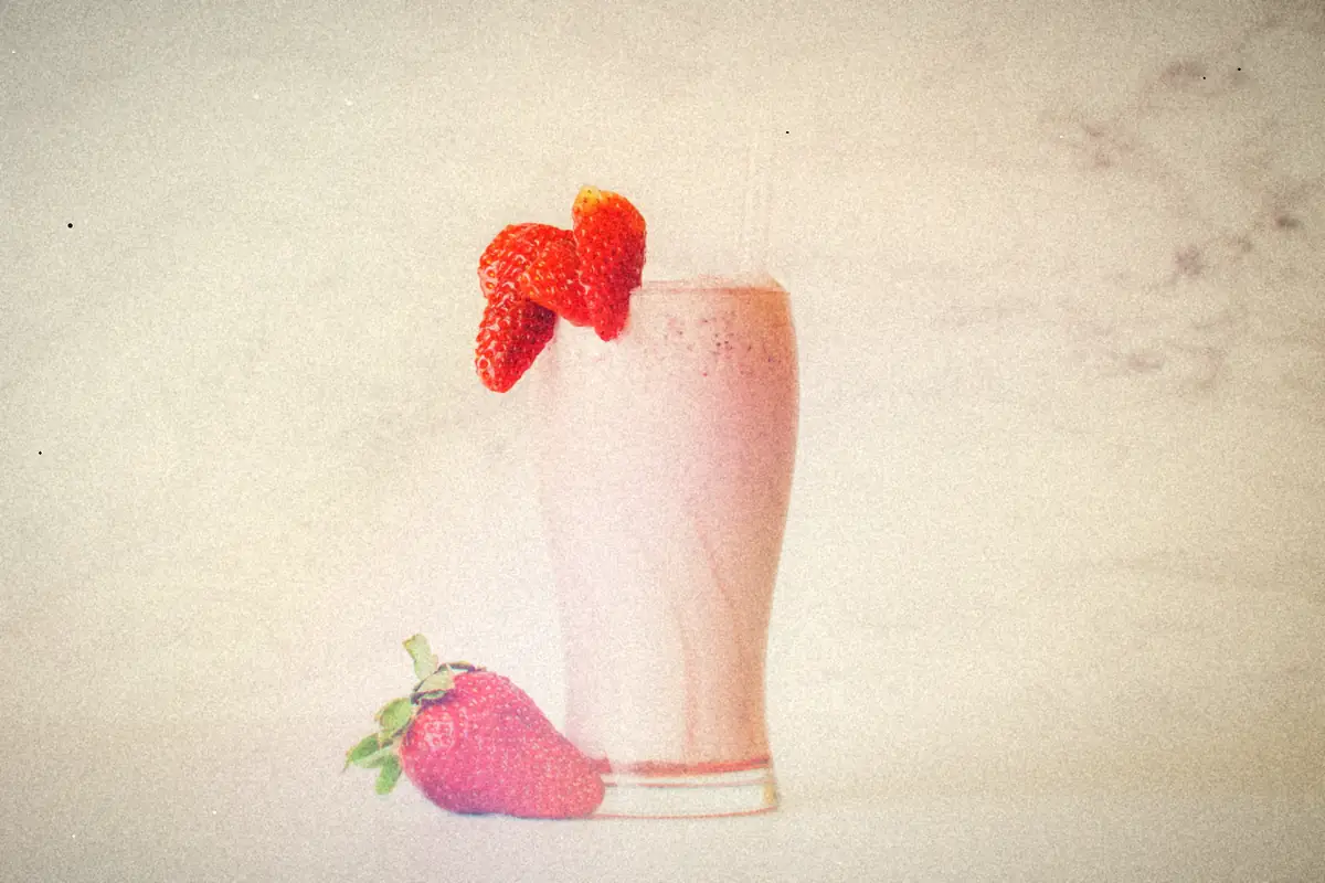A creamy pink strawberry shake in a tall glass with a straw and fresh strawberries on a marble background.