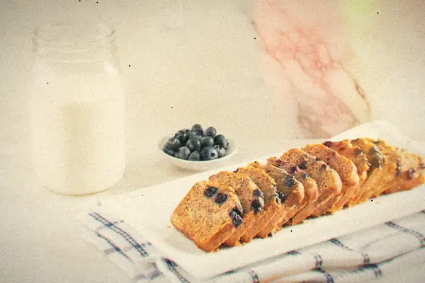 Slices of blueberry bread on a white platter with a jar of milk and a bowl of blueberries.