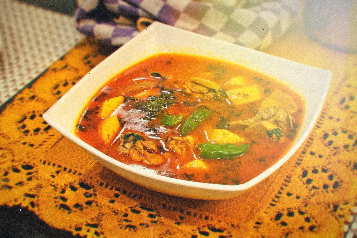 A square white bowl of red-orange chicken soup with potatoes, peppers and herbs.