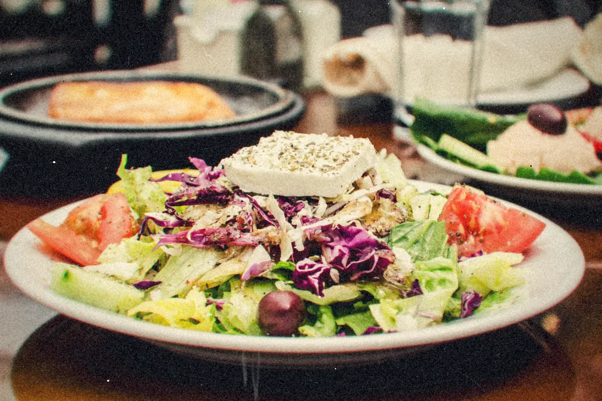 A Greek salad with a slab of feta, tomatoes, cucumber, olives and red cabbage on a white plate.