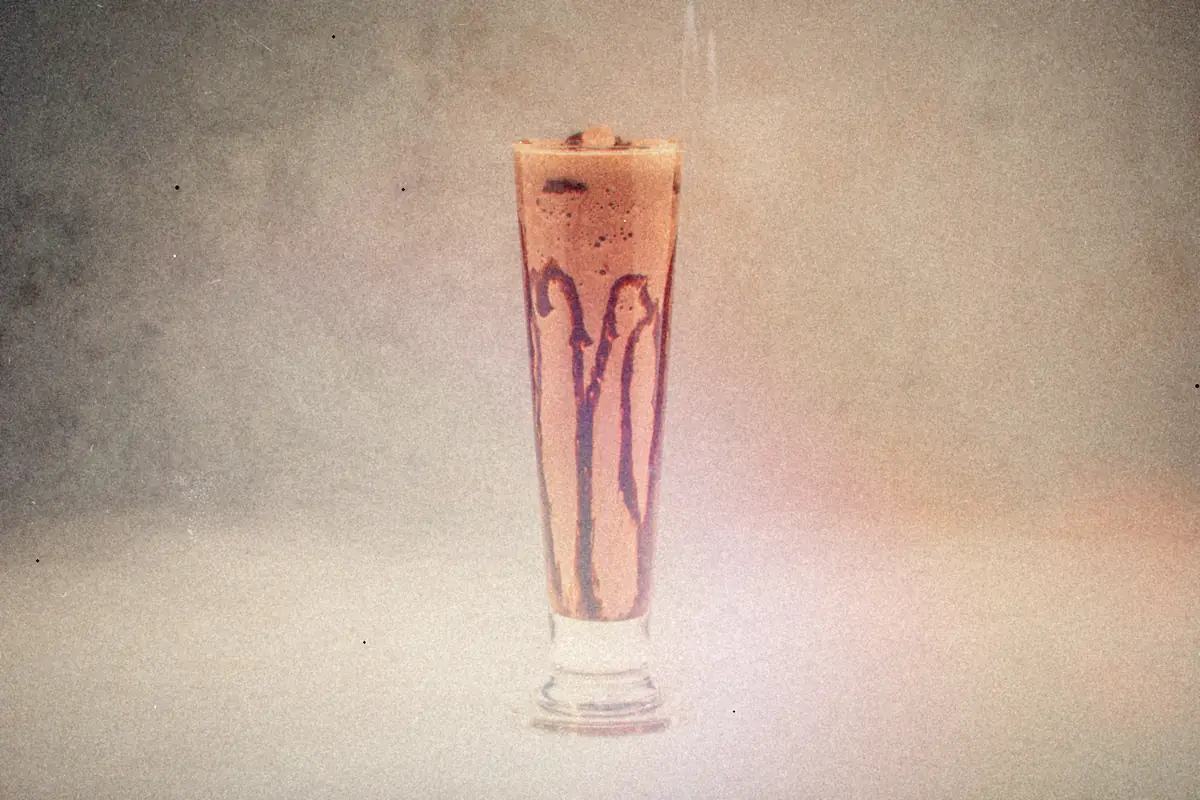 A chocolate shake with chocolate drizzle inside a tall glass on a gray background.