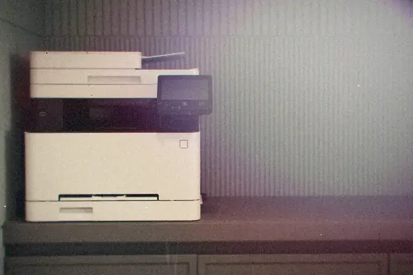 A white multifunction office printer with a small touchscreen on a gray cabinet.