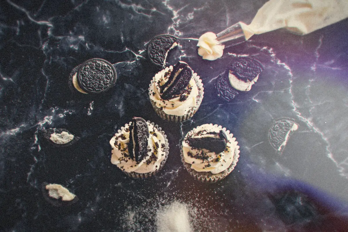 Cookies and cream cupcakes topped with frosting and chocolate sandwich cookies on a dark marble counter.