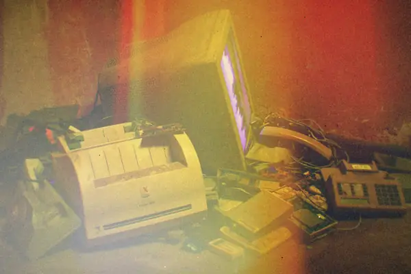 A pile of old electronics, including a printer and a computer monitor, in a dim basement.