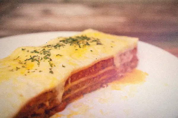A slice of layered meat lasagna with melted cheese and herbs on a white plate.