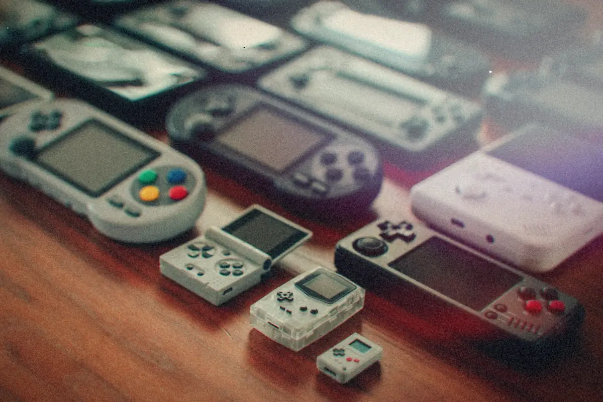 A wooden table covered in retro-style emulation handhelds of different shapes and sizes, from tiny keychain units to wide-screen models.