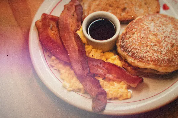 A plate of pancakes, bacon strips and scrambled eggs with a cup of syrup on a wooden table.
