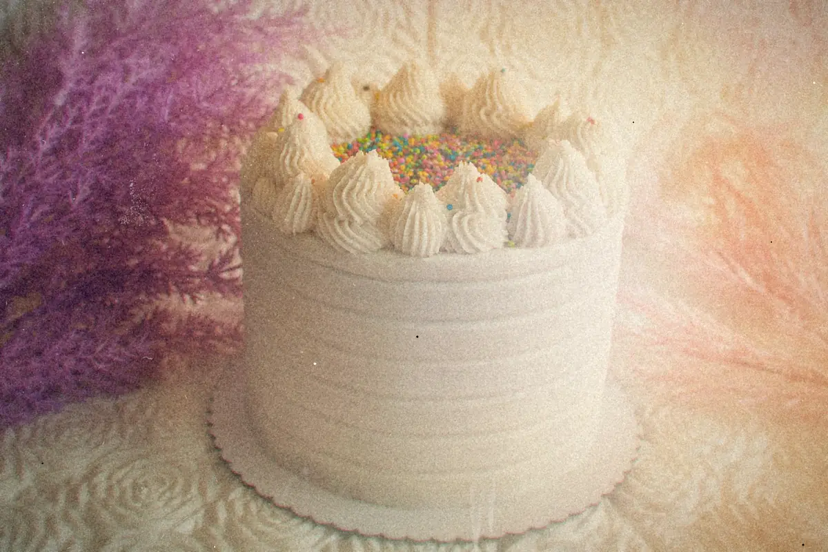 A tall white frosted layer cake topped with piped cream swirls and sprinkles.