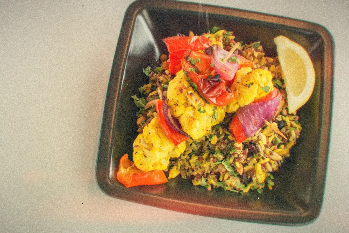 Roasted cauliflower, red peppers and red onion over herbed grains with a lemon wedge in a dark square dish.