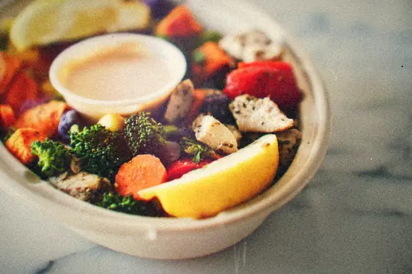 A bowl of seasoned chicken with roasted vegetables, broccoli and lemon wedges, with a cup of dressing.