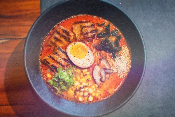 A bowl of spicy ramen with grilled chicken, a soft-boiled egg, corn, mushrooms and scallions.