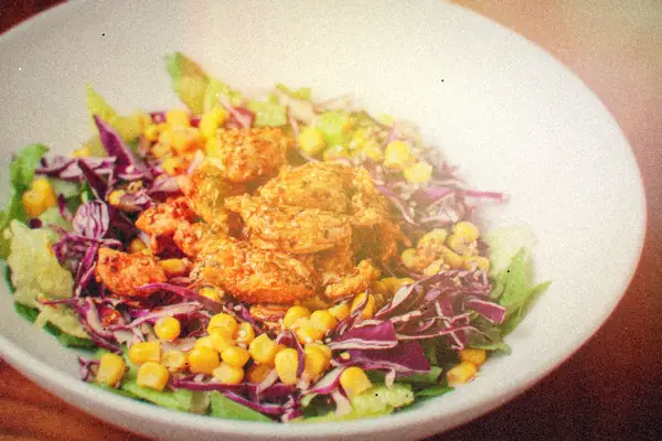 A bowl of spiced chicken over lettuce with red cabbage and sweet corn.