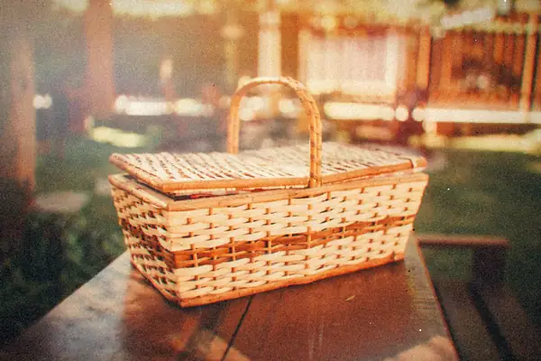 A wicker picnic basket on a wooden table in a sunny backyard with a wooden fence.