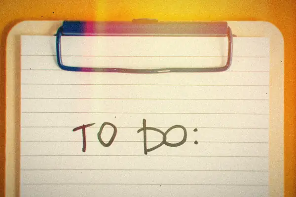 A clipboard holding lined paper with a handwritten to-do heading on an orange surface.
