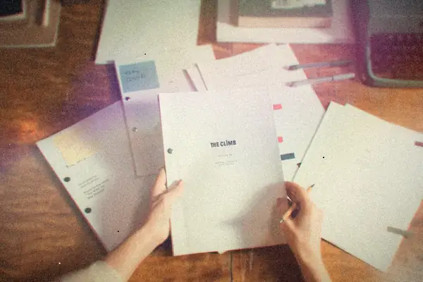 Hands holding a printed manuscript over loose pages and a typewriter on a wooden desk.
