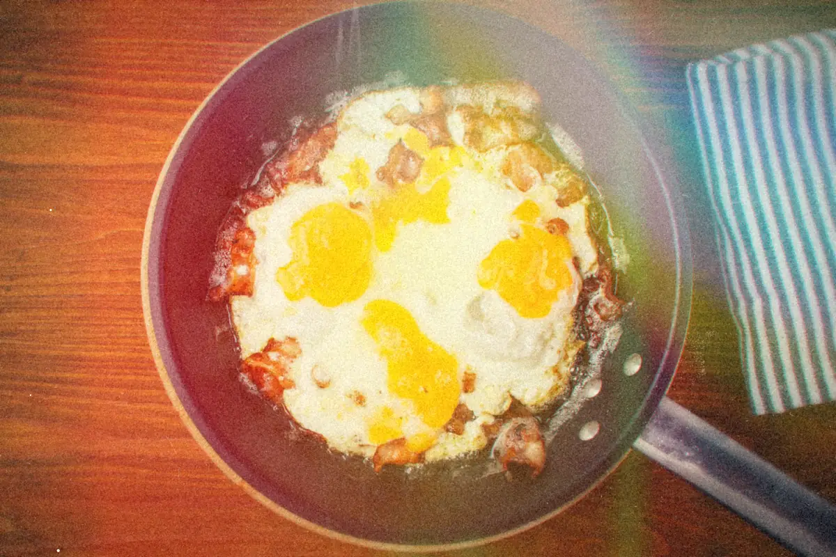 Bacon and fried eggs cooking together in a black skillet on a wooden table.