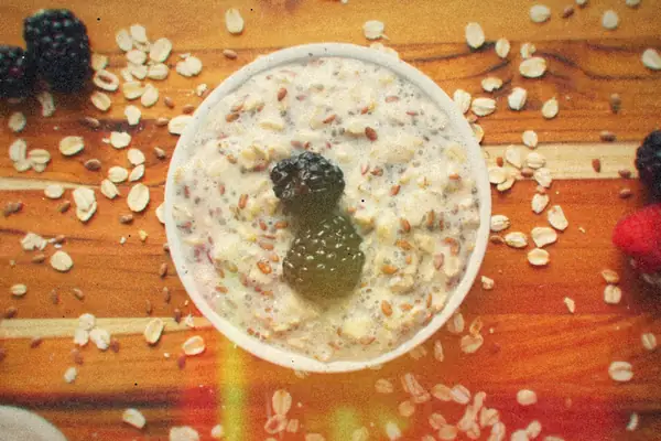 A white bowl of creamy overnight oats with chia seeds, topped with two blackberries, on a wooden table scattered with oats.