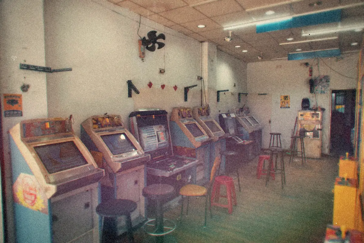 A row of upright arcade cabinets with stools lined up along the wall of a small, brightly lit arcade room.