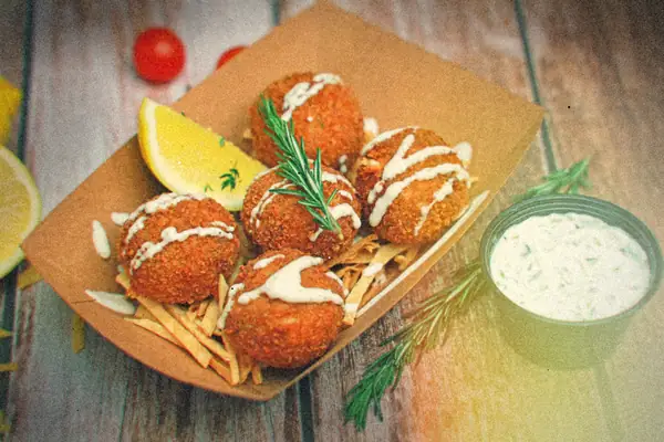 Crispy golden seafood cakes drizzled with sauce in a paper tray with lemon wedges, rosemary and tartar sauce.