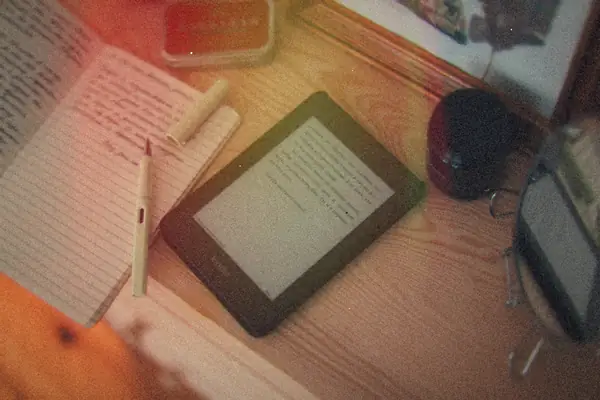 An e-reader lying on a wooden desk next to an open notebook and a pen.