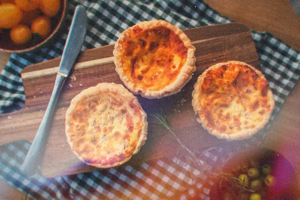 Three baked mini egg quiches on a wooden board beside yellow cherry tomatoes and olives.