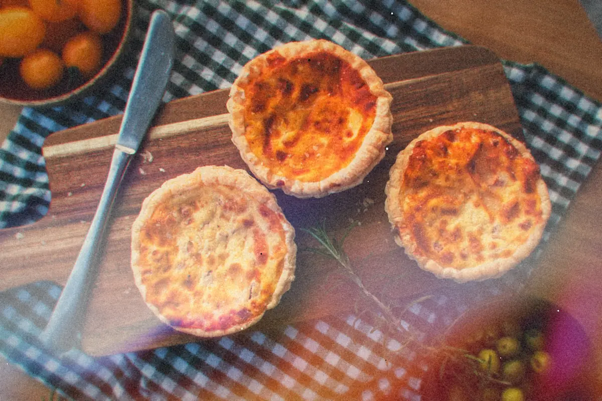 Three baked mini egg quiches on a wooden board beside yellow cherry tomatoes and olives.