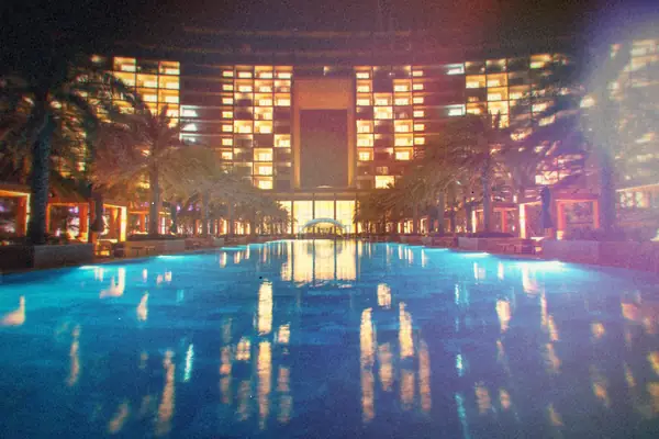 A long swimming pool lit up at night, lined with palm trees.