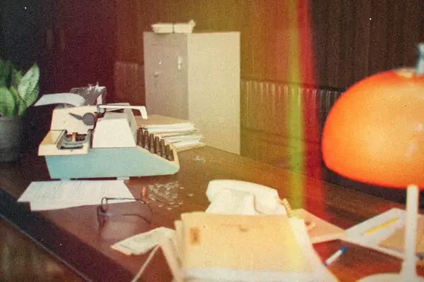 An old office desk with a typewriter, a desk phone, papers and an orange lamp.
