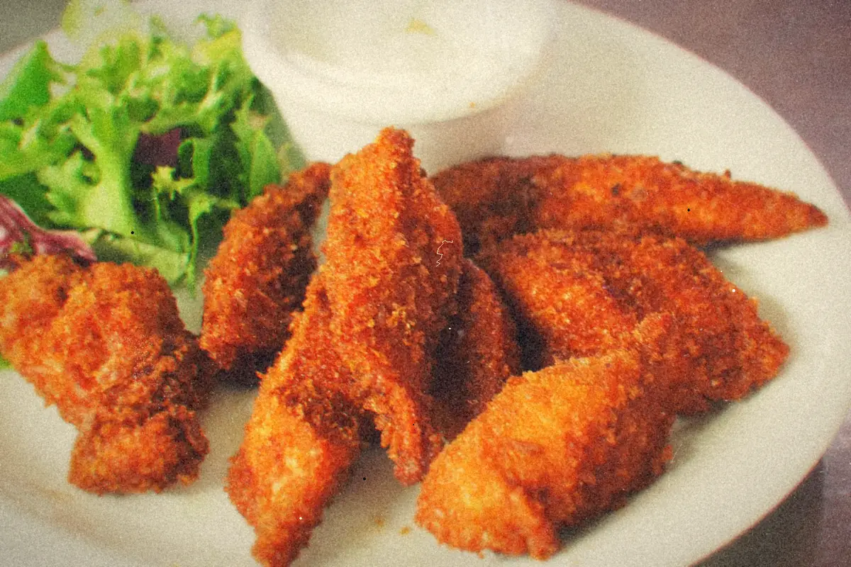 A plate of crispy, deep-golden fried chicken tenders with a side salad and a cup of dipping sauce.