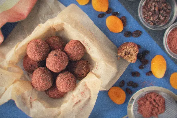 Homemade energy bites rolled in raspberry powder, piled on parchment next to dried fruit and cacao nibs.