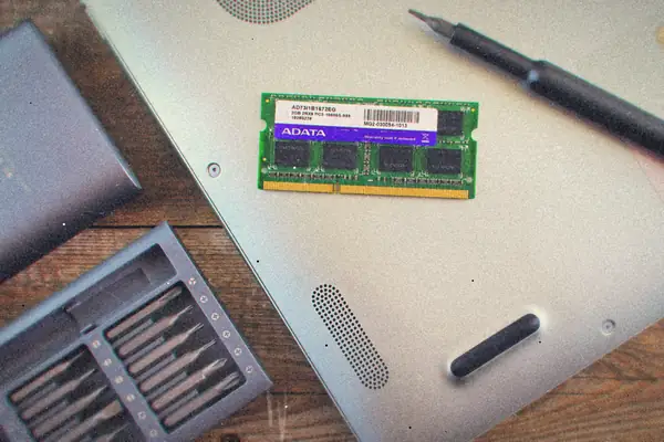 An open laptop underside with a RAM module, a screwdriver and a precision bit set laid out on a desk.