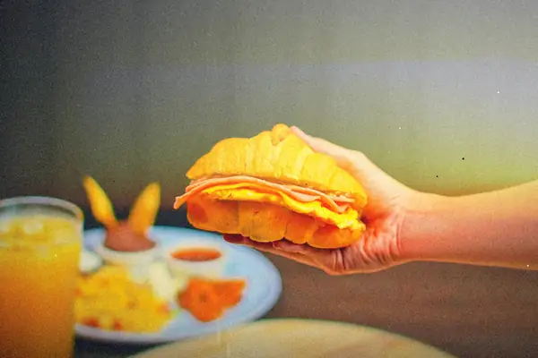 A hand holds a croissant breakfast sandwich with egg and ham over a plate of eggs and a glass of orange juice.