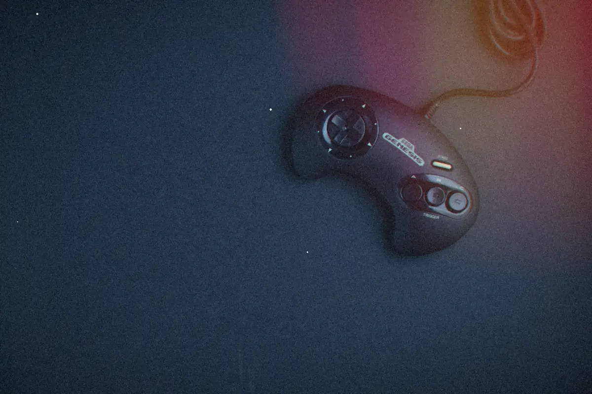A black three-button Sega Genesis controller lying on a dark navy background, its cord trailing off the top of the frame.