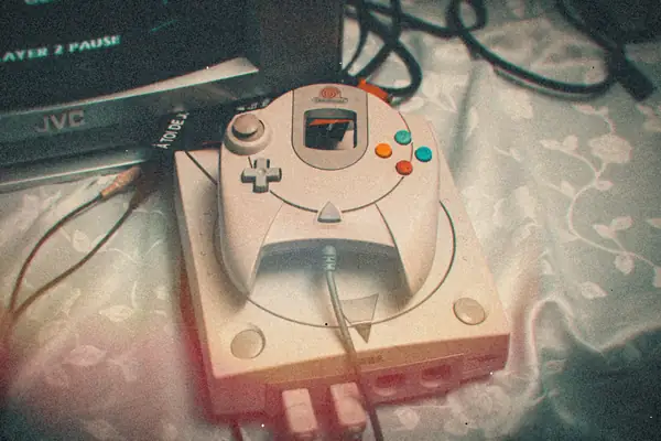 A white Sega Dreamcast with its controller resting on top, cables plugged in, beside an old CRT television.