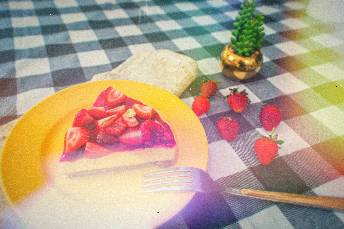 A slice of strawberry-topped cheesecake on a yellow plate with fresh strawberries on a checkered tablecloth.