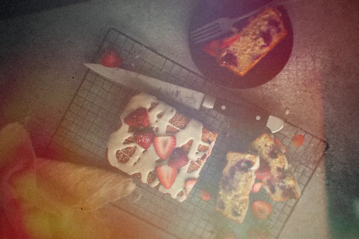 A glazed strawberry loaf cake with fresh strawberries and cut slices on a wire cooling rack.