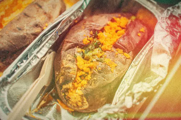 A split baked sweet potato topped with shredded cheese and scallions in a foil tray.