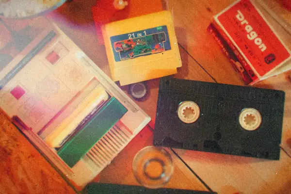 A Famicom-style console with a cartridge in the slot, surrounded by more 8-bit cartridges, a VHS tape and a drink on a wooden table.